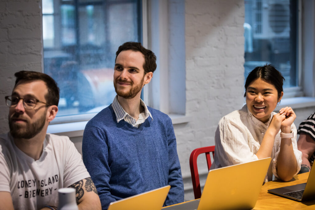 Three colleagues sitting at a table, working on laptops and smiling at the camera.